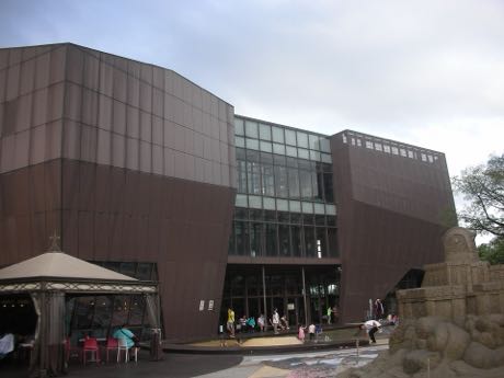 深褐色金屬外牆的大型場館，中央為玻璃帷幕入口；左前方有白色遮陽傘與桌椅，右側是一組大型沙雕作品，入口處有遊客進出。
