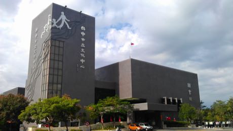 深灰色量體的文化中心建築，左側外牆有大型人形與線條圖案及中文館名；建築前有樹木、旗桿與停放的車輛。