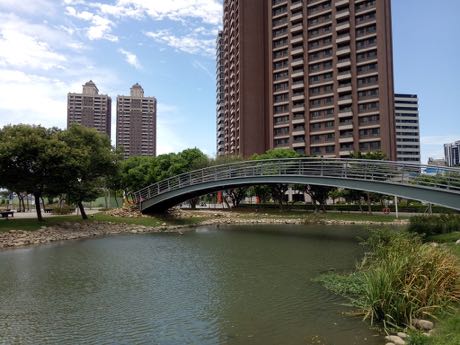 公園水池上的白色拱橋，橋面有金屬欄杆；兩岸為草坡與樹木，水邊長有蘆葦，後方是多棟高層住宅大樓，天空藍白相間。
