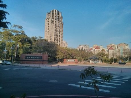 街道對面的公園入口，紅色矮牆上有園名字樣，後方是樹木與一座涼亭；遠處可見高層住宅大樓，前景為馬路與行人穿越道。