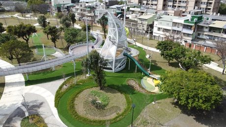 從空中俯瞰八德楓樹腳公園，白色長頸鹿造型遊戲塔旁有綠色管狀溜滑梯，一條高架棧道環繞在草地與樹林間。