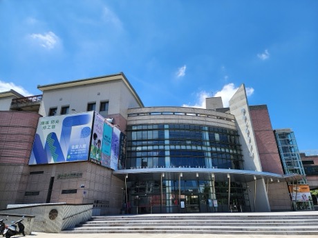 藍天下的博物館建築，正面為弧形玻璃帷幕與雨遮，左側外牆掛著展覽布幕；建築前有一段階梯與廣場。
