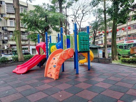 福德公園的紅黃藍綠四色塑膠組合遊具與兩道滑梯，設於紅灰相間的橡膠地磚上，後方是掛著紅燈籠的街道。