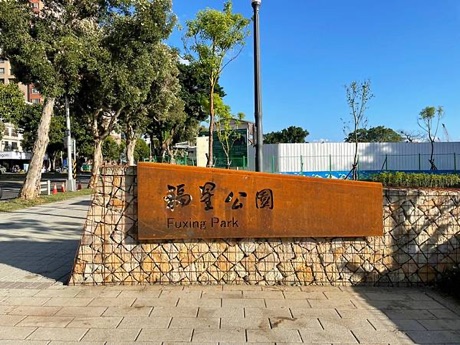 福星公園入口的耐候鋼板名牌寫著「福星公園 Fuxing Park」，立在石籠牆上，後方是樹木與藍天。