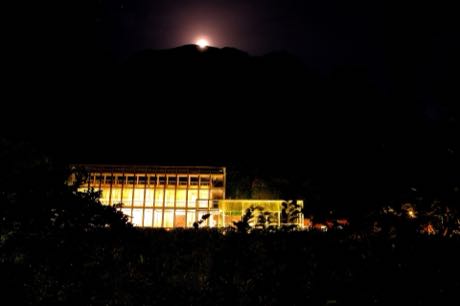 夜間的山區建築，長方形量體的玻璃立面透出暖黃燈光；背後為黑色山影，山頭上方有一輪明亮月光，前景是深色樹叢。