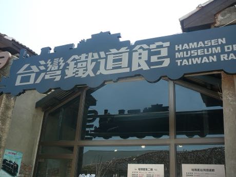 館舍入口上方的深色造型招牌，鏤刻「台灣鐵道館」中文與英文館名，招牌下緣為蒸汽機車輪廓；下方是玻璃門窗與解說牌。