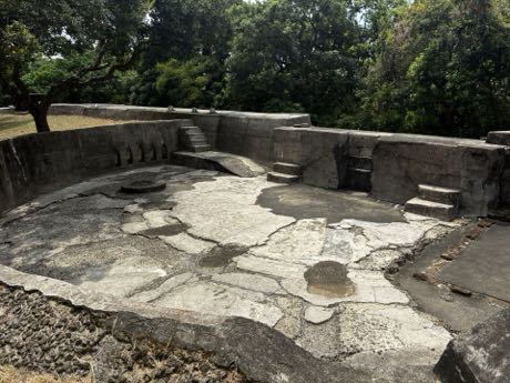 砲台遺構的混凝土砲座平台，中央地面龜裂、積有水漬，周圍為弧形厚牆與數段石階；牆外是樹林。