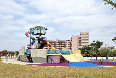 健康綠洲公園的大船造型遊戲場，有紅藍相間的旋轉管狀溜滑梯、瞭望塔、攀爬網與攀岩坡，前方是草地。