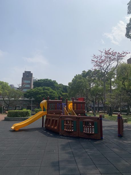景華公園的船身造型木構遊戲組，附綠色遊戲面板與黃色滑梯，後方有一株盛開的粉色花樹。