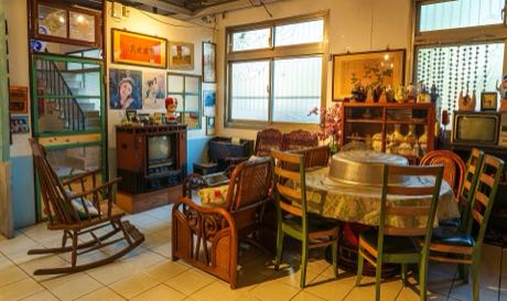 復原的舊式住宅室內，擺放木製搖椅、藤製沙發與圓桌餐椅；牆面掛有舊海報與相框，角落有老式電視機與玻璃櫥櫃。