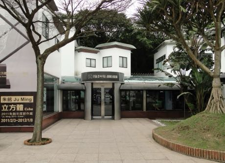 美術館入口建築，白色牆體與深色屋簷，門上有館名標示；左側立有一面展覽海報看板，前方是樹木與弧形花台。