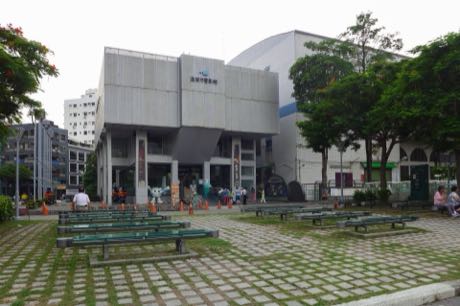 灰色清水模外牆的館舍建築，入口處掛有直式布幕；前方廣場鋪石磚並設有多張長椅，數名民眾在門口與廣場活動，四周有樹木。