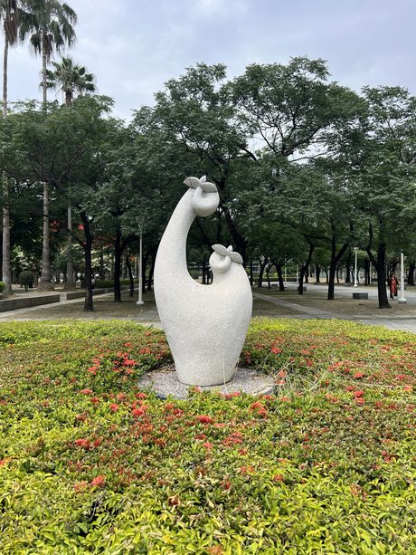 高雄勞工公園草地上的白色抽象雕塑，周圍種滿紅花與矮灌木，後方是成排大樹。