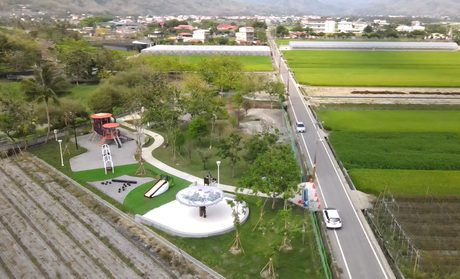 空拍美濃湖畔特色遊戲場，綠地上有溜滑梯和遊戲設施，旁邊是道路與稻田。