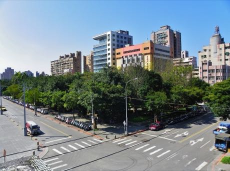從高處俯瞰街角，中央為一片被樹林覆蓋的公園綠地；周圍是多線道路、行人穿越道與停放的機車，背景為市區大樓。