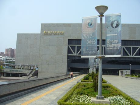 大型博物館建築的側面，米色石材外牆與方正量體，牆上有中文館名字樣；前方有兩面直式展覽布旗、燈柱與花圃，一名行人走在步道上。