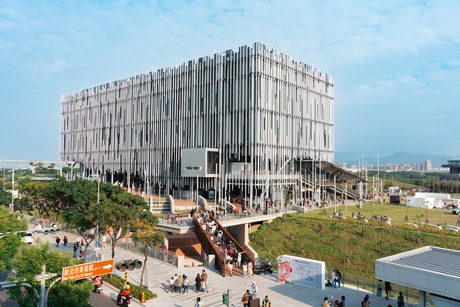 新北市美術館的長方形量體外覆密集細長金屬立柱，右側連接大片綠色草坡與戶外園區。