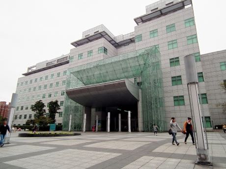 多層樓的灰白色圖書館建築，入口為綠色玻璃帷幕與挑高雨庇；前方是寬闊的石板廣場，數名行人經過，旁有燈柱與植栽。