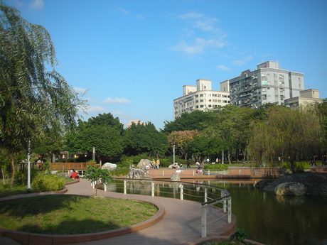 八二三紀念公園的水池與弧形紅磚步道，白色欄杆的木棧道跨過水面，岸邊有垂柳與置石。