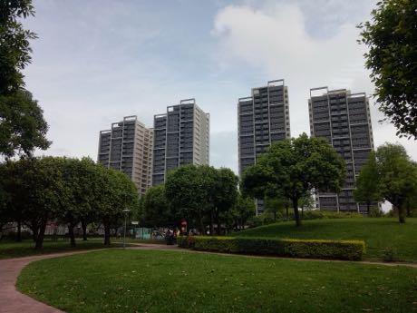公園草地與步道，前景為修剪過的草皮與灌木；後方有數棟高層住宅大樓與成排樹木，天空多雲。