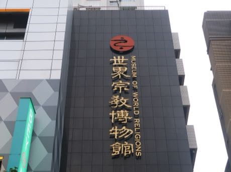 由下往上拍攝的大樓外牆，深色牆面上有直式金色立體字「世界宗教博物館」與英文館名，上方為紅色圓形標誌。