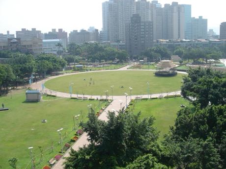 從高處俯瞰的都市公園，中央為橢圓形大草坪與環繞的步道、路燈；四周種有樹木與花圃，遠處是密集的高層住宅大樓。