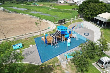 空拍柳營櫻花滯洪池公園，藍色船造型遊戲場與鞦韆位在滯洪池旁，後方是長滿草的滯洪池與稻田。