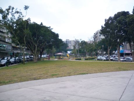 社區公園的草地與水泥步道，中央遠處有一處帶頂棚的遊戲設施區；四周是大樹、停放的汽車與住宅大樓。
