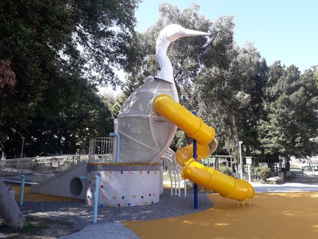 蘆洲三民公園的鷺鷥造型遊戲塔，白色鷺鷥頭頸立於細網編織的塔身上，連接兩段黃色管狀滑梯，旁有攀岩立面與橘色防護鋪面。