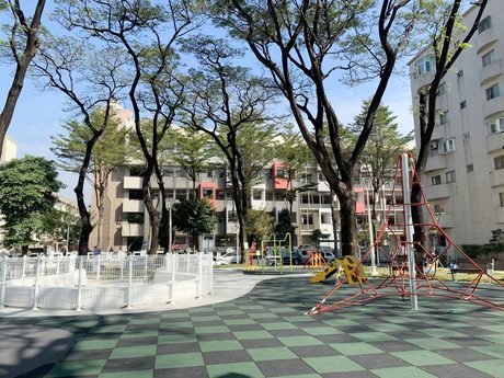 汕頭公園的綠黑棋盤格地墊上有紅色攀爬網塔、長頸鹿滑梯和白色圍欄沙坑，四周是大樹。