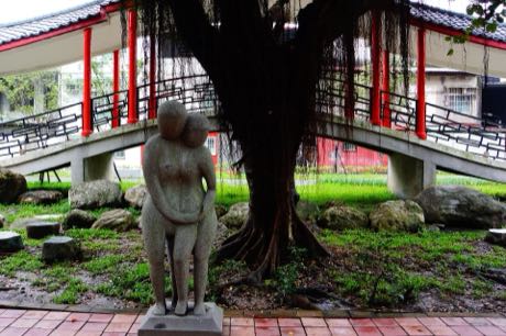 公園中的石雕作品，為一對相依的人形石像；後方是一棵氣根垂落的大榕樹與紅柱白頂的廊道，地面有草地與散置石塊。