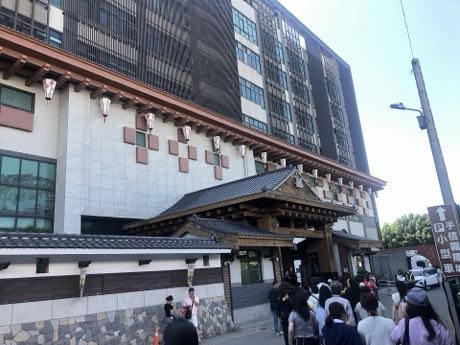 日式風格的建築入口，黑瓦屋簷與木格柵門廊，上方外牆有木質裝飾與吊燈；門前有多名遊客排隊，右側為街道與招牌。