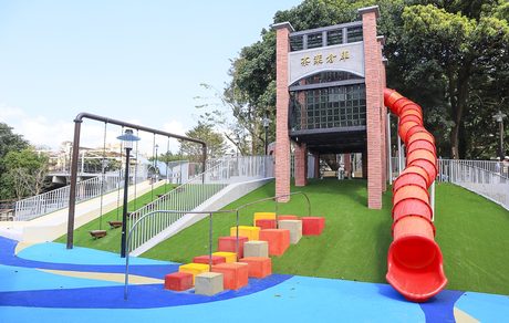 水返腳公園乾式遊戲區，磚柱鋼構的茶葉倉庫造型遊戲塔連接紅色螺旋管狀滑梯，前方有鞦韆、彩色跳樁與藍色波浪鋪面。