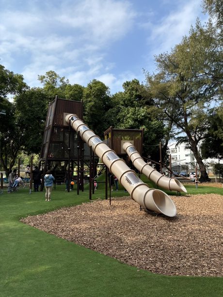 后里森林公園樹蔭下的雙軌不鏽鋼隧道溜滑梯，滑梯出口鋪有木屑軟墊。