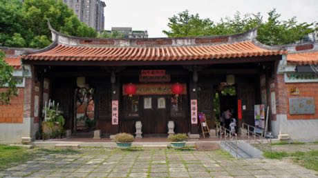 傳統三合院式建築的正廳，紅瓦屋頂與燕尾脊，簷下懸掛紅燈籠與匾額，門旁貼有紅色對聯；前方是石板埕與盆栽，背景有高樓。