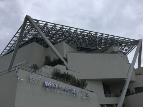 由下往上拍攝的美術館建築，白色幾何量體層層堆疊，上方覆蓋大型白色鋼構格柵屋頂；牆面有中英文館名與「2館」字樣，天空陰灰。