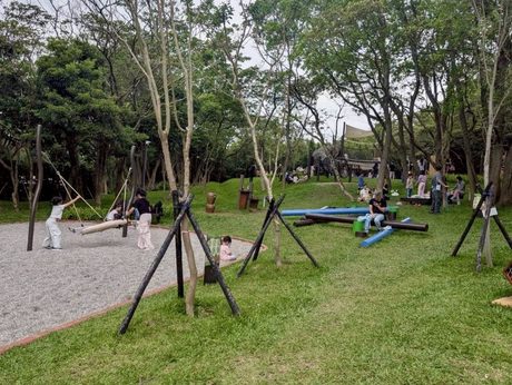 五酒桶山自然遊憩場的林間遊戲區，礫石地上有鳥巢鞦韆和原木平衡木，後方樹林中有遮陽帆下的木造攀爬設施。