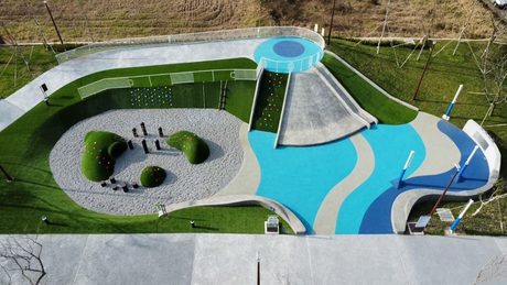 從高處俯瞰新龍公園的共融遊戲場：藍色波浪地墊、大片磨石子溜滑梯、攀岩牆，以及鋪白色碎石、有綠色小山丘的沙坑區。
