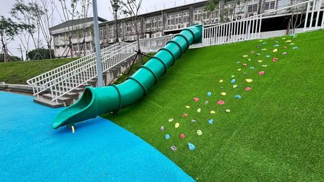 兒22公園的草坡遊戲區，綠色管狀溜滑梯沿坡而下，旁邊草坡上有彩色攀岩點，底下是藍色地墊。