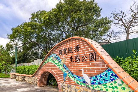 鑊篤陂塘生態公園入口的紅磚拱形園名牆，牆面寫著「鑊篤陂塘生態公園」，下方有白鷺鷥和翠鳥的彩色馬賽克，後方是大樹。