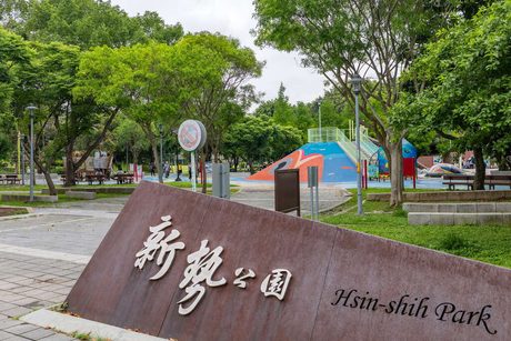 新勢公園入口刻有「新勢公園 Hsin-shih Park」的石牌，後方樹蔭下可見彩色的遊戲場。