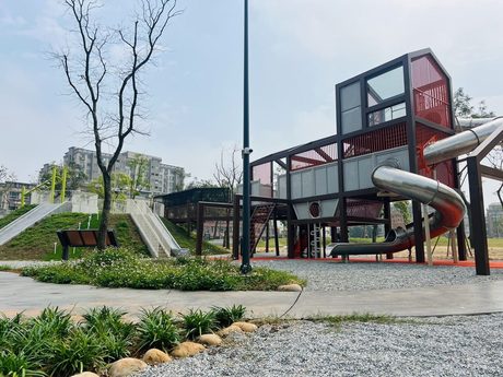 四維兒童公園的眷屋造型遊戲塔，紅色框架屋頂下接著長長的不鏽鋼螺旋溜滑梯，前方是草地和碎石步道。