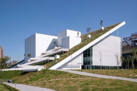 白色量體的現代館舍建築，一道斜向的長坡道自地面延伸至屋頂綠地；建築前為草坡與步道，天空湛藍。