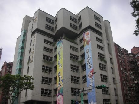 多層樓的白色圖書館建築，立面為規則排列的窗戶與凸出量體；牆面懸掛兩幅直式活動布幔，樓下可見街道與號誌。