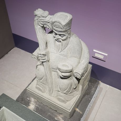 展示中的石雕神像，為一尊長鬚、戴帽、持杖並手捧元寶的坐姿土地公；像身立於雕花石座上，背景為紫色展牆。