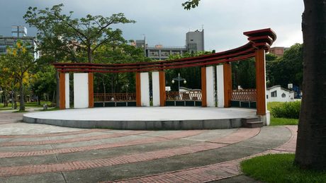 溪北公園的圓形表演舞台，後方是橘柱紅頂的弧形棚架與座位看台，四周為喬木與紅磚鋪面。