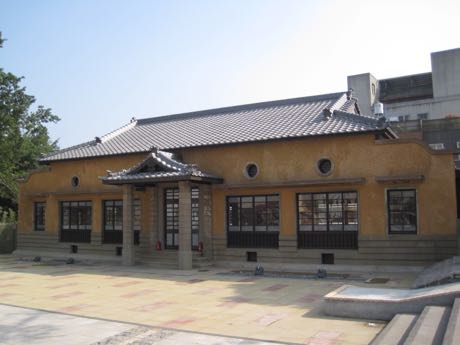 日式木造建築，灰瓦屋頂與土黃色牆面，中央有小型門廊與木格窗，牆上有圓形裝飾；前方為鋪面廣場，天空湛藍。
