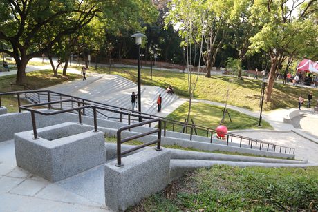祥合公園的無障礙坡道與階梯沿著高低地形而下，四周是大樹和草坡，右側可見沙坑。