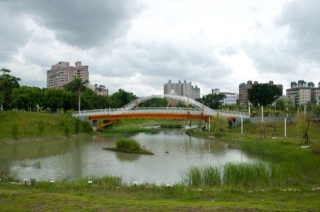 公園滯洪池與跨池的橘白色拱橋；池中有一小片植生島，岸邊長有水草，背景為住宅大樓與樹木，天空多雲。