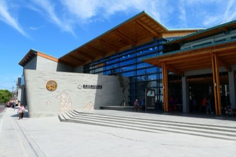 館舍建築，灰色石材牆面搭配木構屋簷與玻璃帷幕，牆上有中文館名與圓形標誌；前方為寬階梯與廣場，數名遊客在門口活動，天空湛藍。